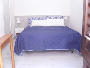 A neatly made bed with a blue bedspread and white pillows, a wooden nightstand with a lamp, and a white drawer unit against a light-colored wall.
