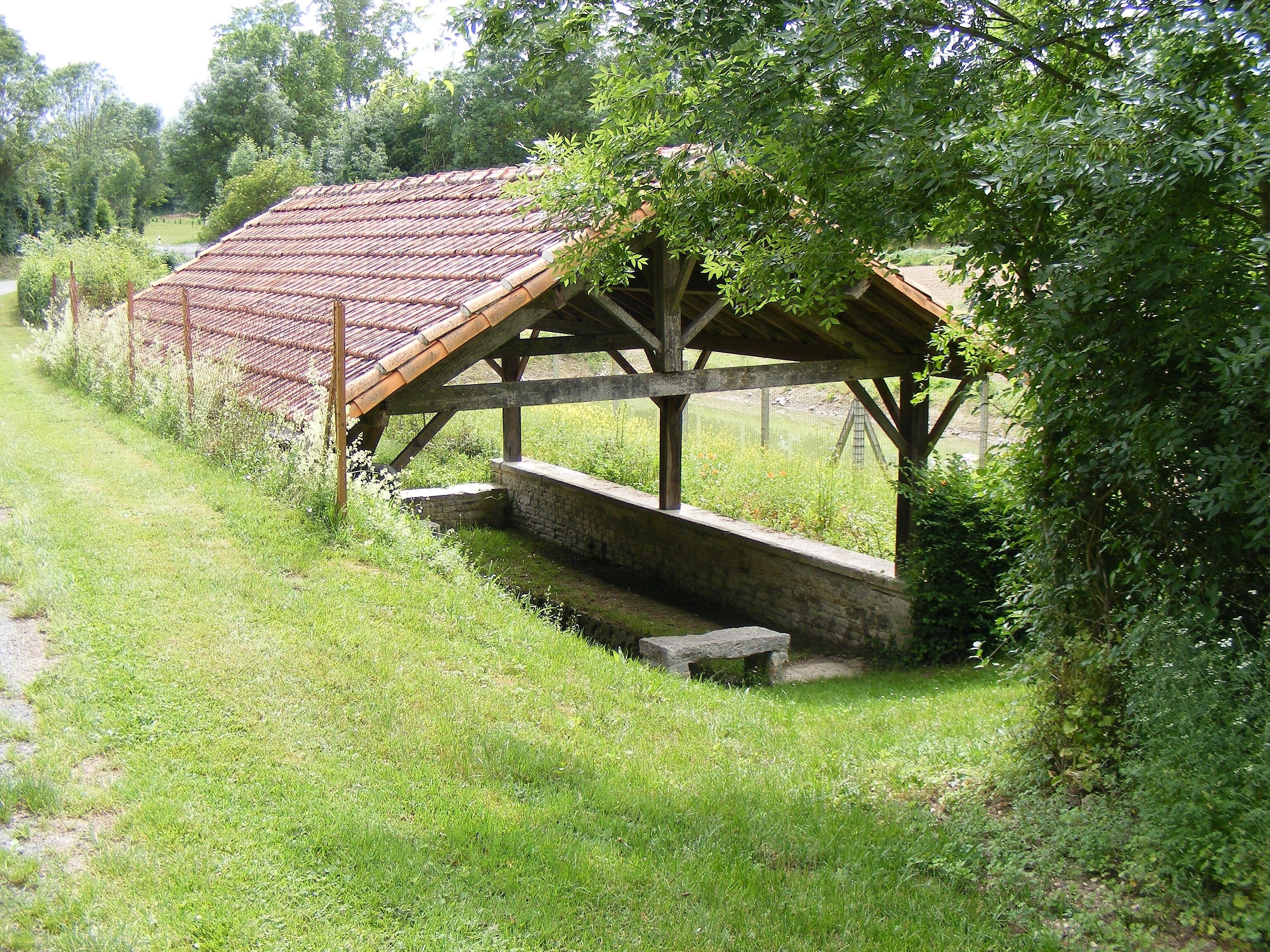 Une construction rustique en bois, dotée d’un toit en tuiles, entourée d’une végétation luxuriante et d’une zone herbeuse.