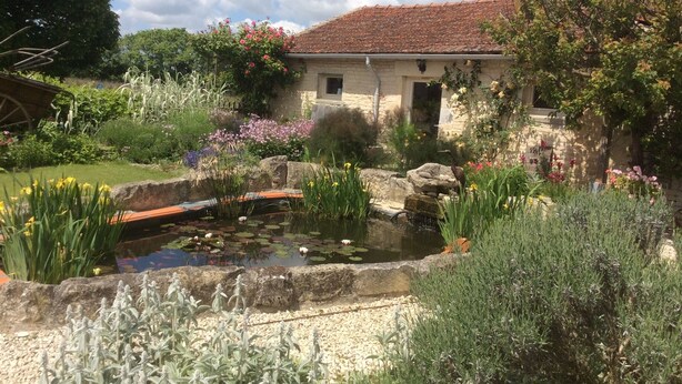 A rustic stone building with a tiled roof, surrounded by a garden with a pond, various plants, and flowers. - Cosy, pet friendly cottage for 2 in pretty countryside (Crézières)