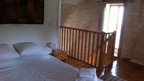 A bedroom with a large bed, a wooden nightstand, a small table, and a view of a stone wall and balcony. - Cosy, pet friendly cottage for 2 in pretty countryside (Crézières)