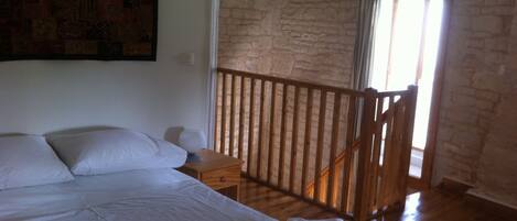 A bedroom with a large bed, a wooden nightstand, a small table, and a view of a stone wall and balcony.