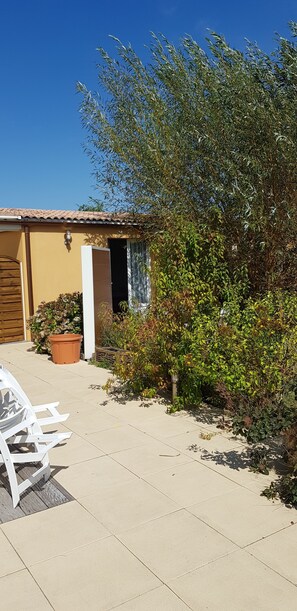 Uma casa com um pátio ladrilhado, uma porta de madeira e um jardim com diversas plantas e uma cadeira branca.