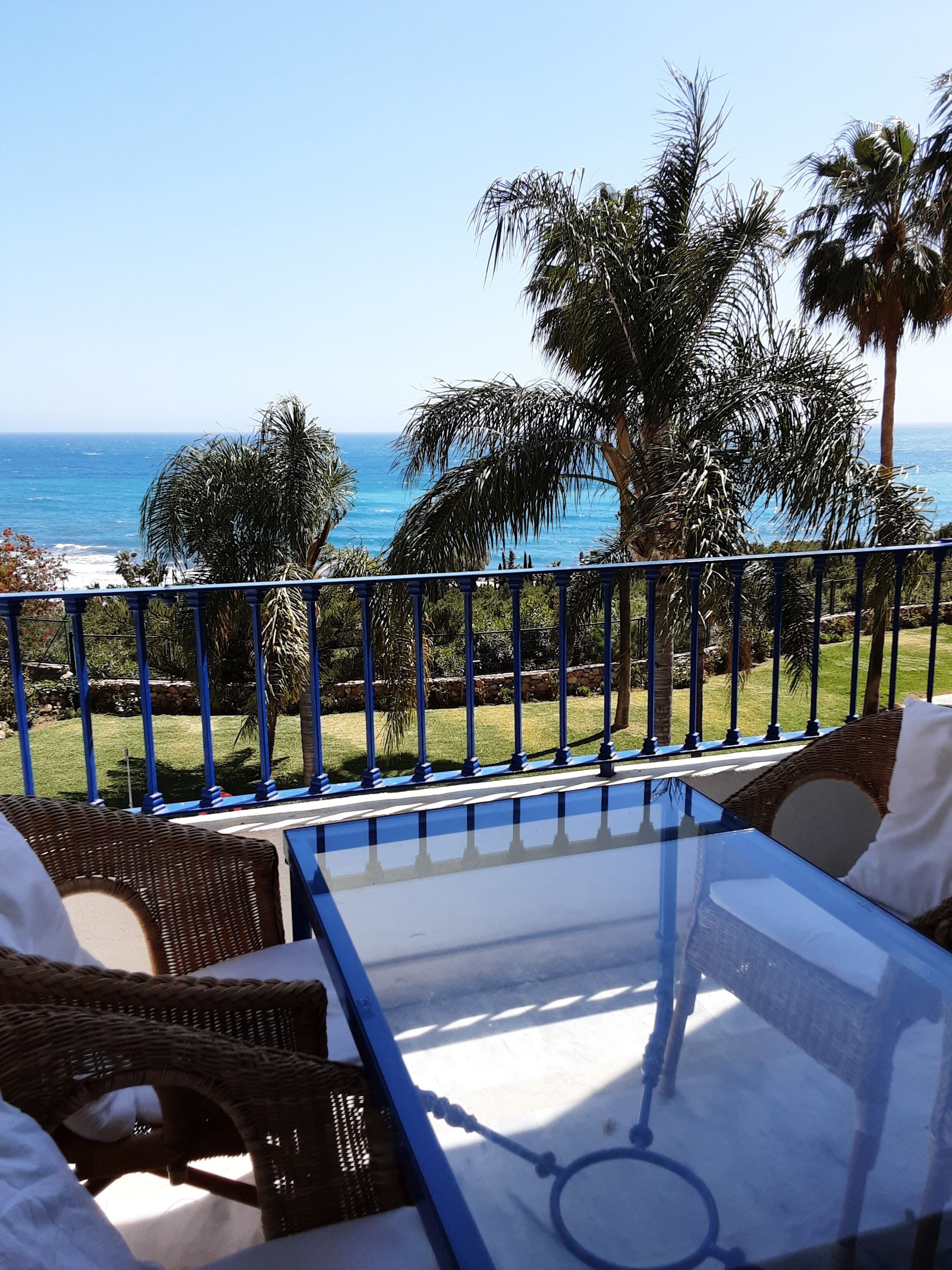 Un balcon avec une table en verre et des chaises en osier, donnant sur une plage et des palmiers.