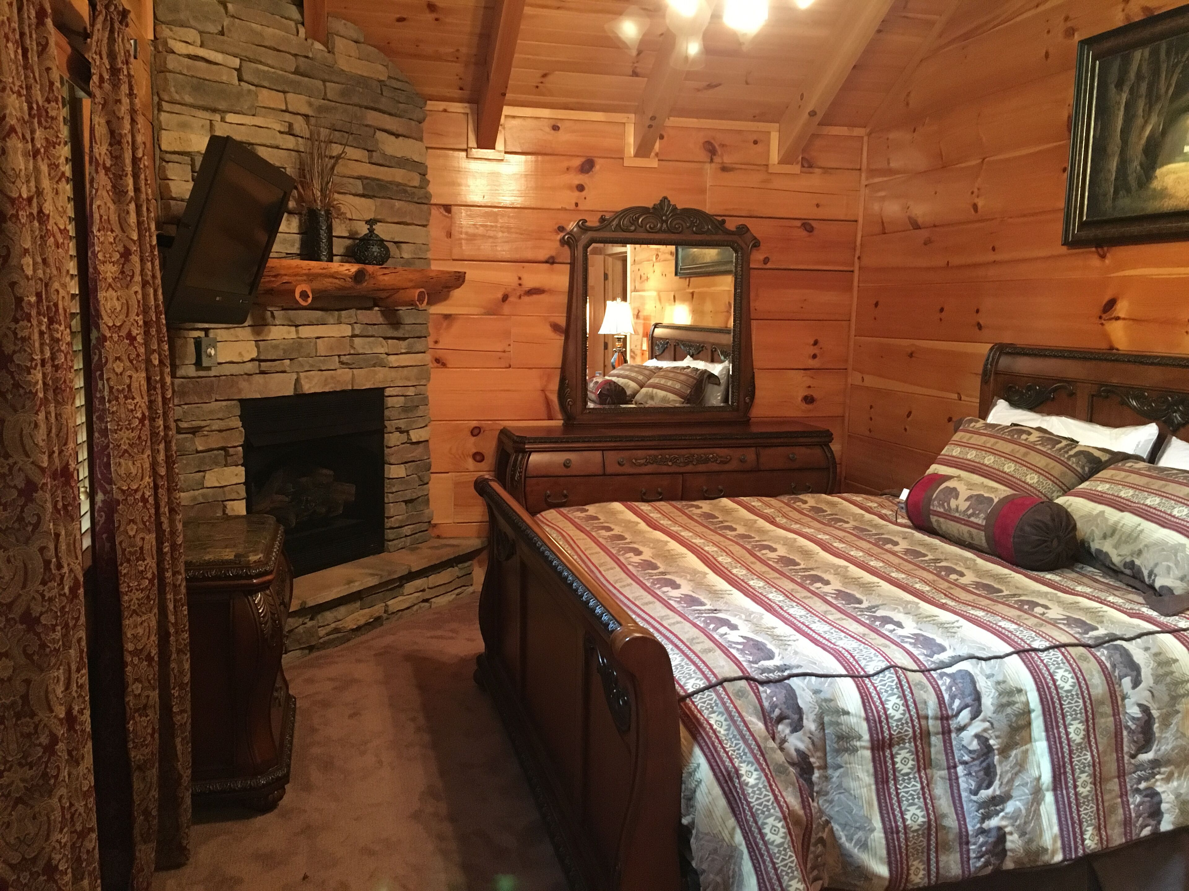 A cozy log cabin bedroom with a stone fireplace, a wooden bed with patterned bedding, a dresser, and a mirror reflecting the room.