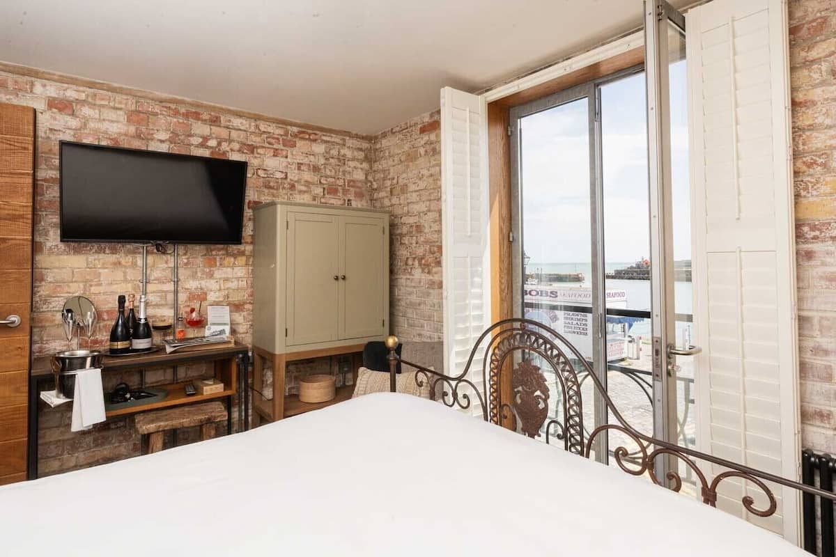 A bedroom with a brick wall, a bed, a television, a cabinet, and a window with a view of the sea.