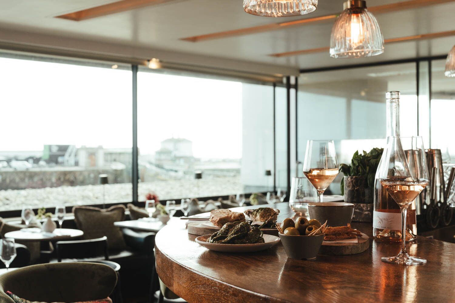 A restaurant with a view of a cityscape, featuring a wooden bar with wine glasses, a bottle of wine, and a bowl of olives.