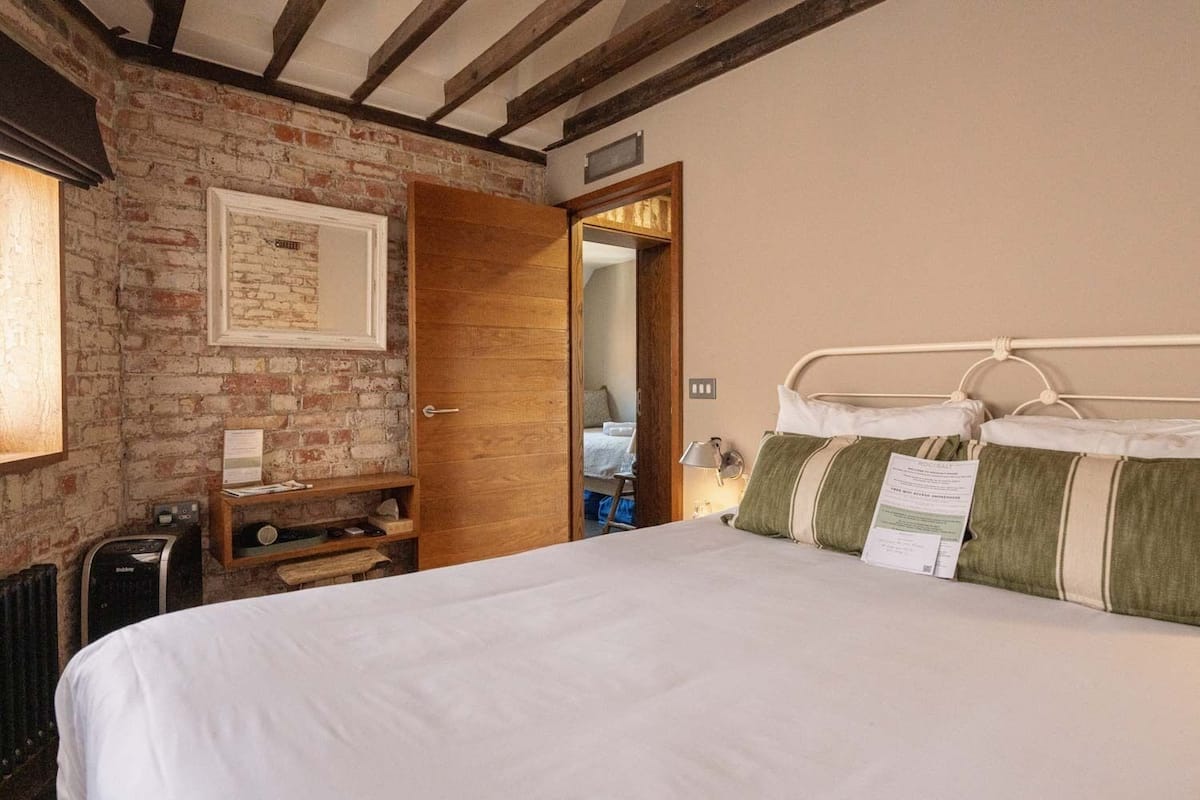 A bedroom with a brick wall, a wooden door, a bed with white bedding, and a bedside table with a lamp.