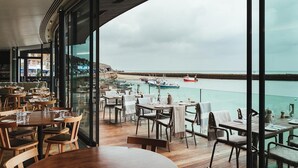 Un restaurante con mesas y sillas dispuestas en una terraza de madera, con vista a un puerto con barcos y una costa rocosa.