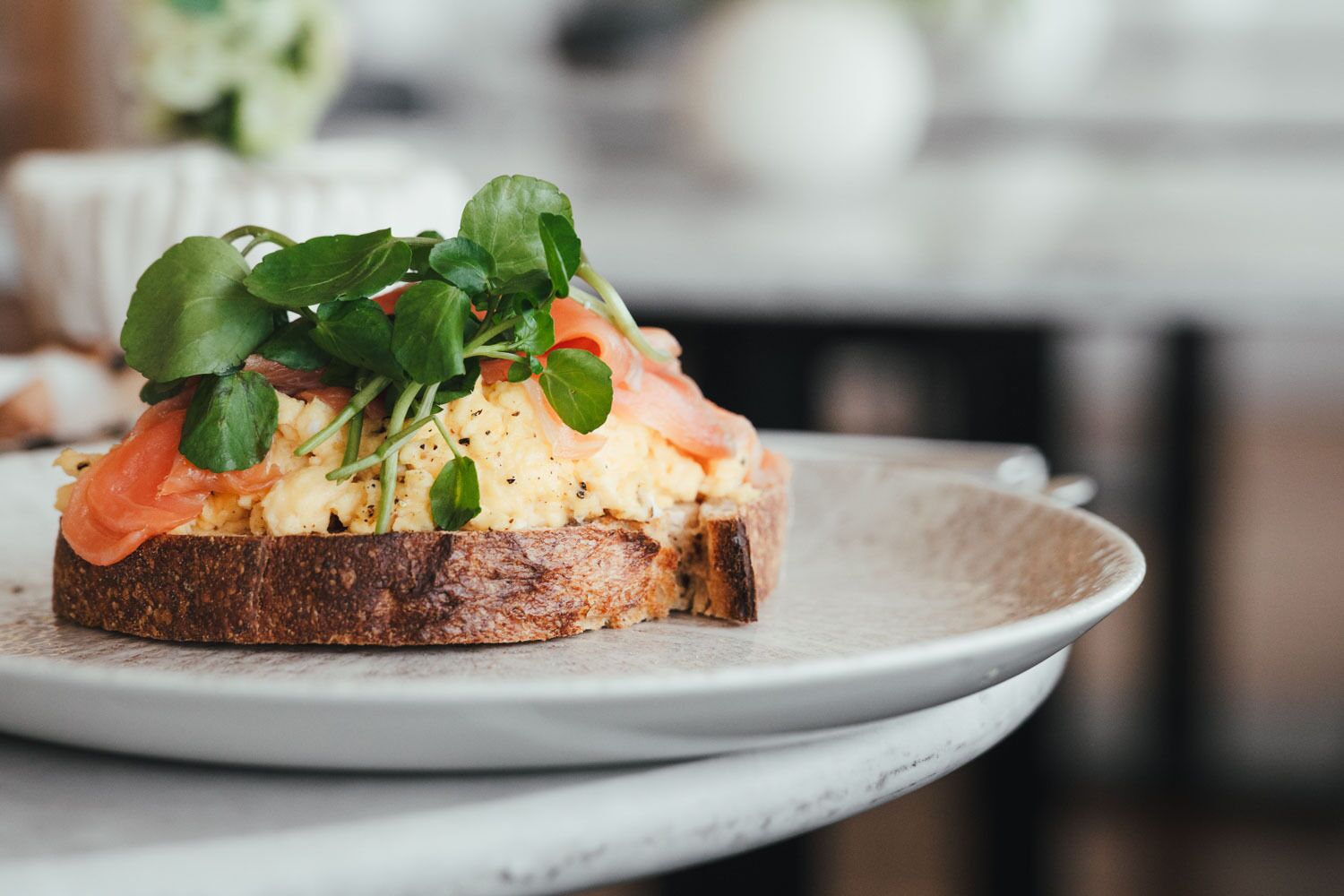 A plate with a sandwich topped with greens and salmon, served on a white ceramic plate.