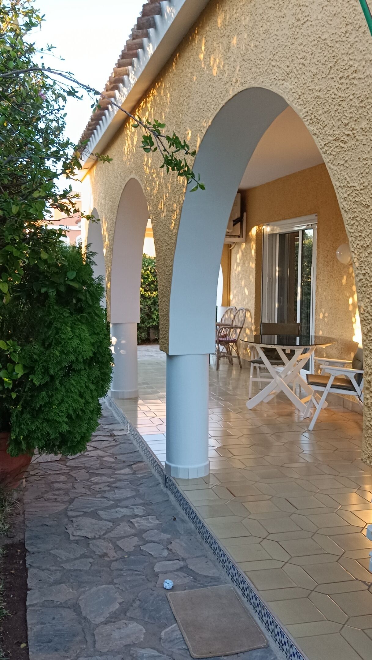 A covered outdoor patio with stone flooring, white pillars, and a seating area with chairs and a table.