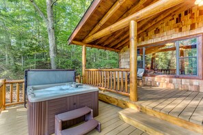 A wooden deck with a hot tub, wooden railing, and a covered seating area with a view of the forest.