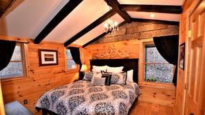 Un dormitorio de cabaña de madera con una cama grande, una araña y dos ventanas con cortinas.