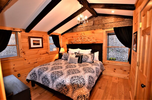 A wooden cabin bedroom with a large bed, a chandelier, and two windows with curtains.