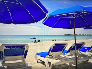 Uma praia com guarda-sóis azuis, cadeiras de praia brancas e um píer ao fundo.