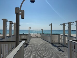 Um calçadão à beira-mar com piso em grade de metal, guarda-corpos brancos e vista clara para o oceano.