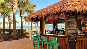 Eine Strandbar mit einem Reetdach, einem Neon-Schild mit der Aufschrift 'COLD BEER' und einer Speisekarte, die Cocktails auflistet.