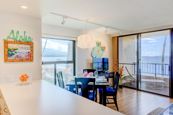 A modern dining area with a large window, a table set for two, and a view of the ocean. - Beautiful Oceanfront Corner Unit (Kihei)