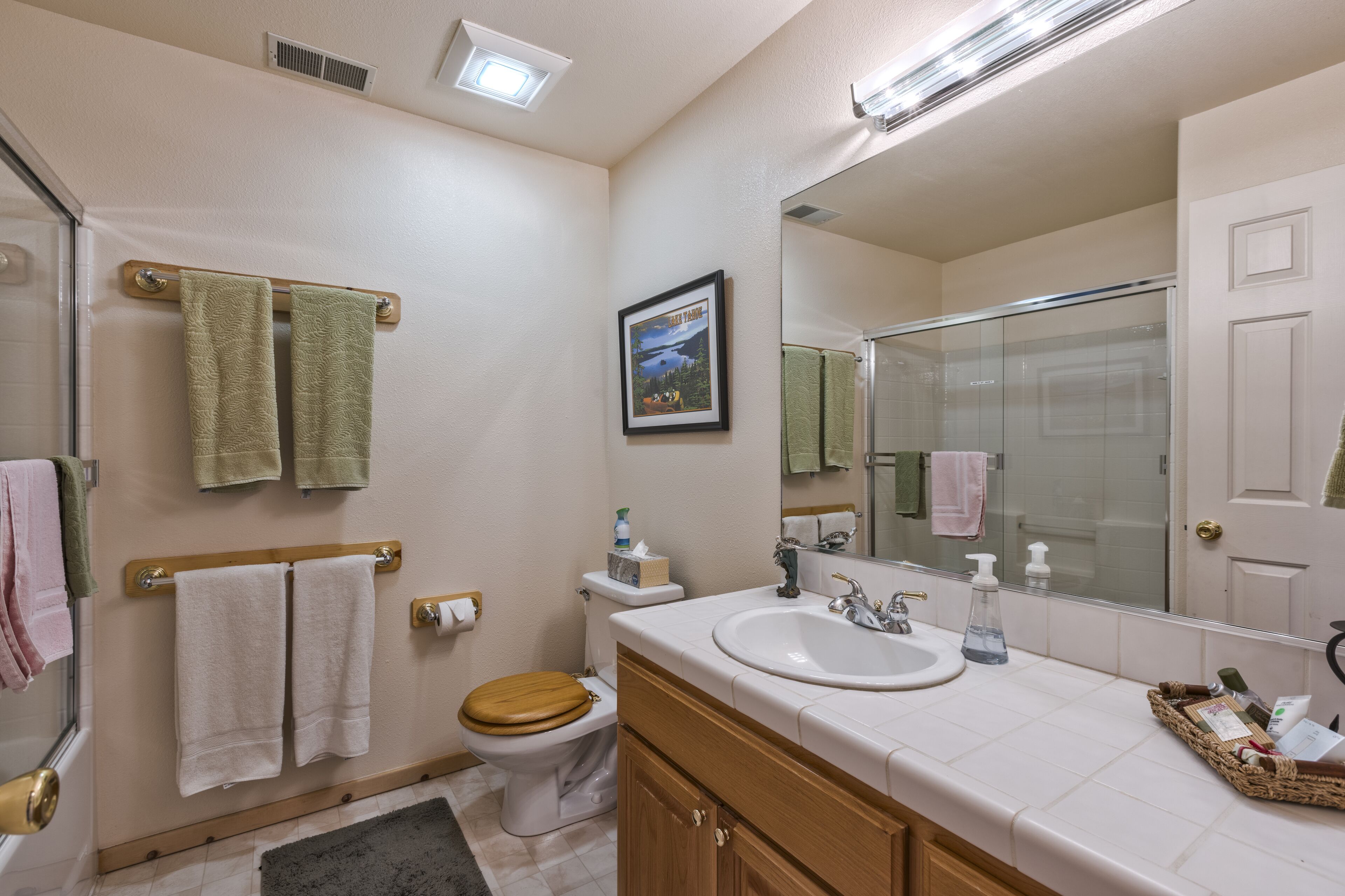 A bathroom with a toilet, sink, and shower. There are towels hanging on the wall and a framed picture above the toilet.