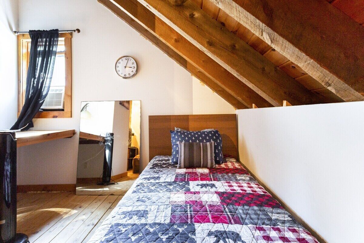 Ein Schlafzimmer mit Holzbalkendecke, einem Bett mit Quilzudecke, einer Uhr und einem Fenster mit Vorhängen.