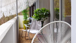 Een balkon met een witte parasol, twee witte rieten stoelen, een tafeltje met een roze kaars en bloempotten.
