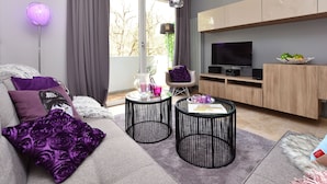 Een moderne woonkamer met een grijze bank, paarse kussens, een ronde salontafel en een televisie op een houten kast.