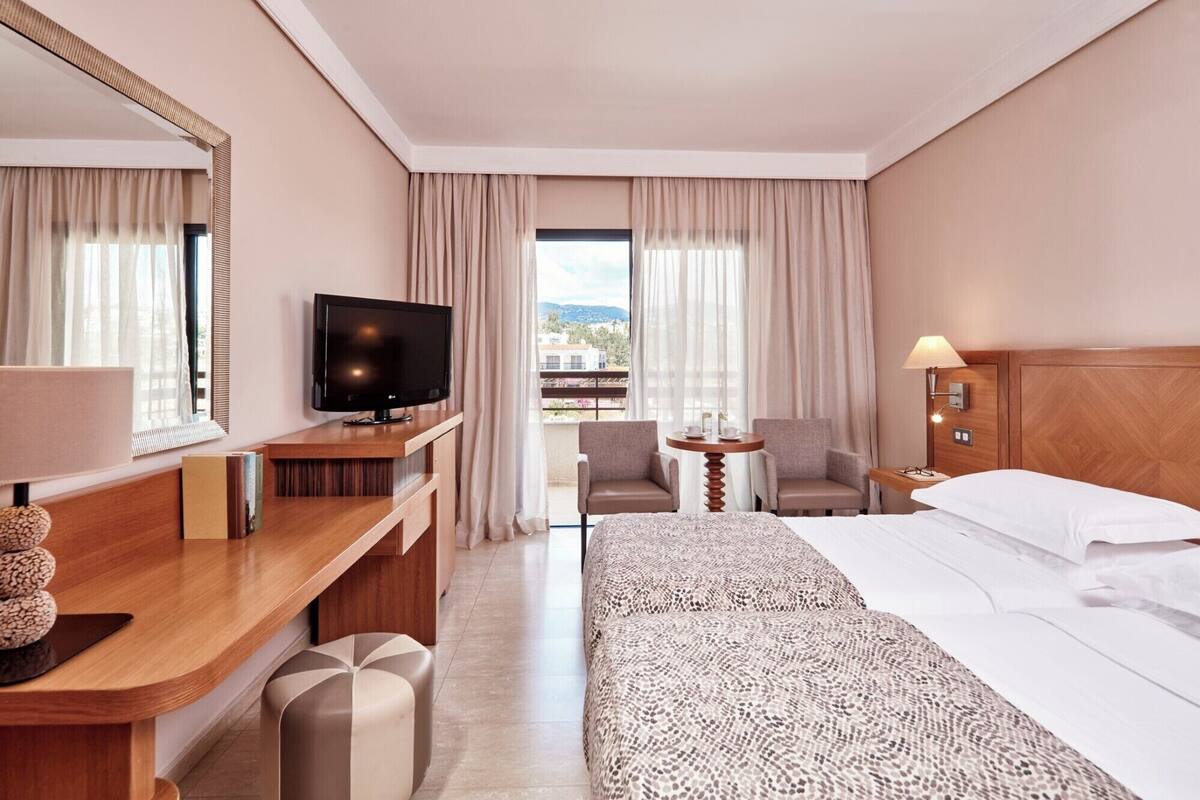 A hotel room with a bed, a desk with a TV, a chair, a small table, and a lamp.
