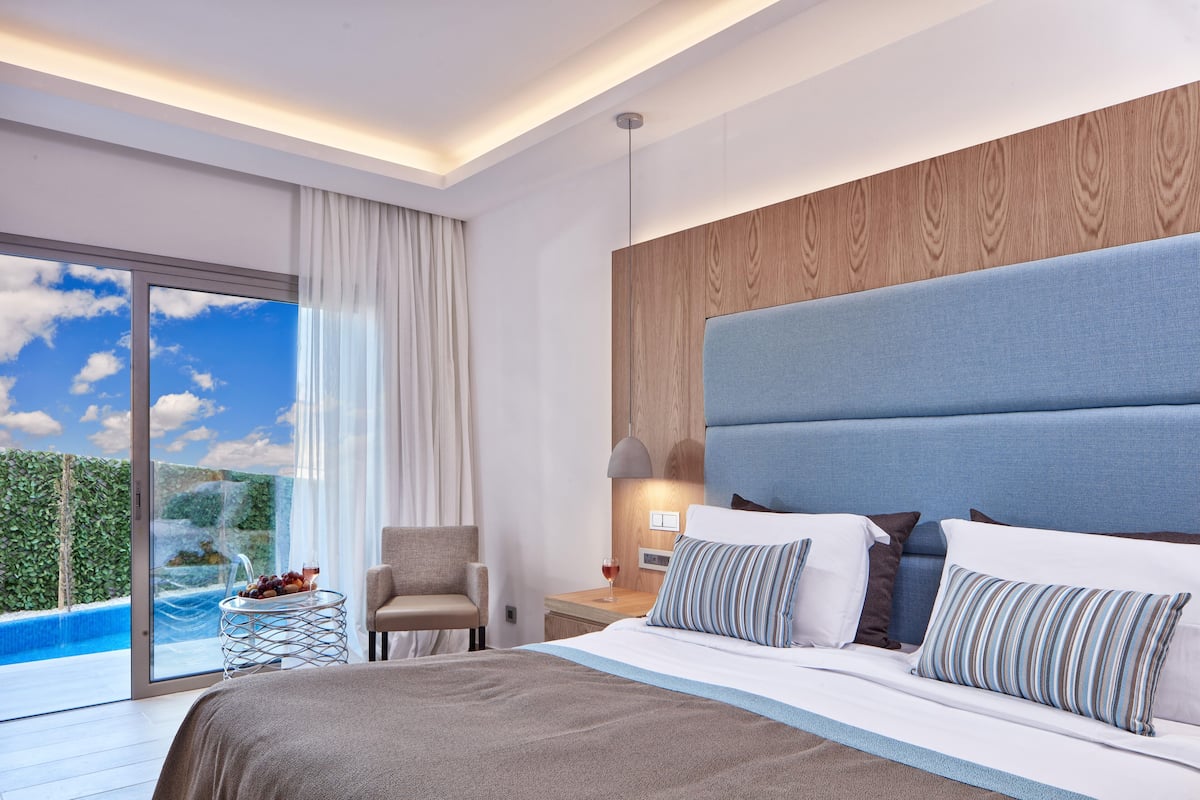 A modern hotel room with a large bed, a seating area, a nightstand, and a view of a pool.