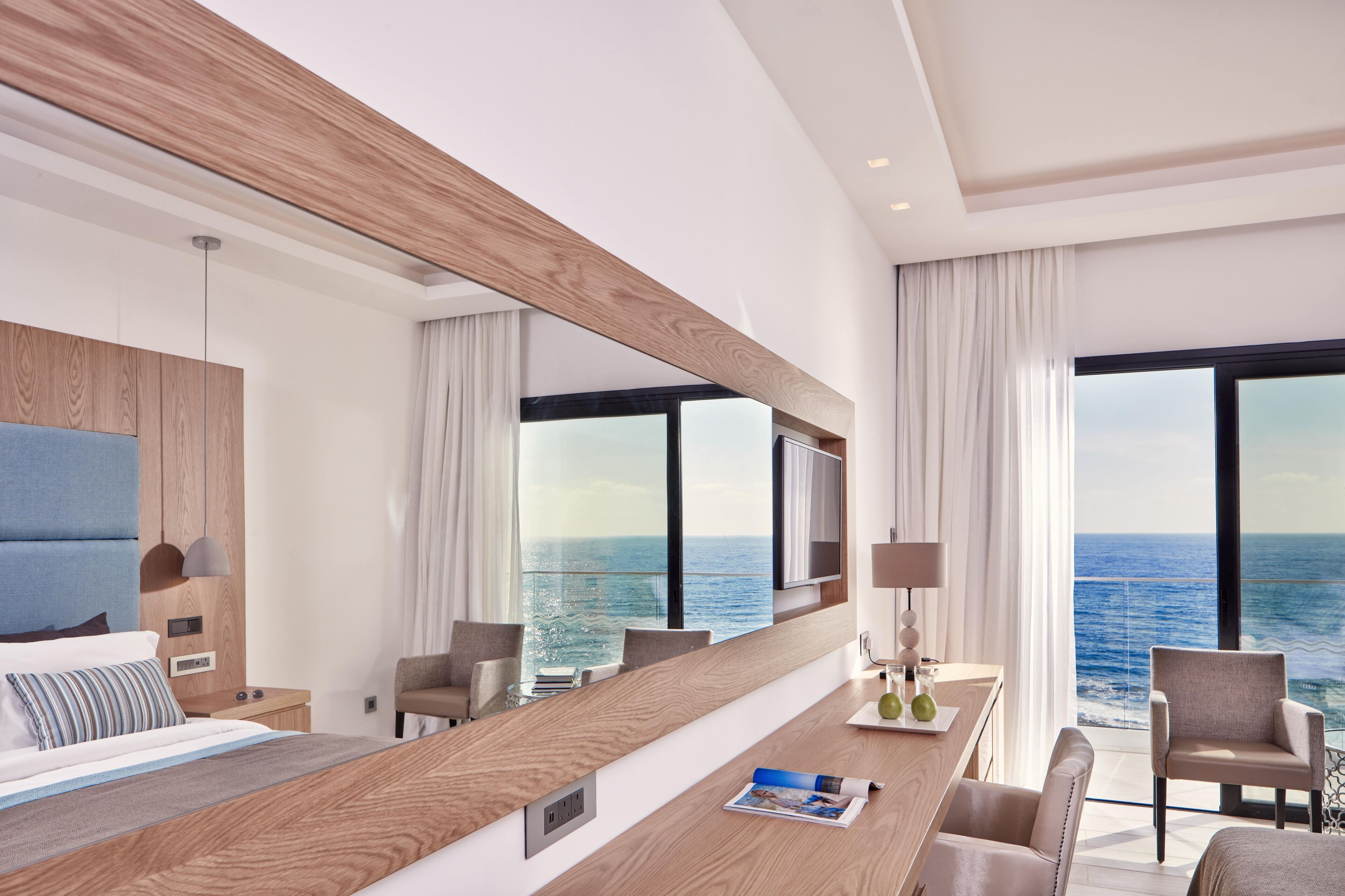 Une chambre d’hôtel moderne avec un grand lit, un bureau en bois, une télévision et une vue sur l’océan.