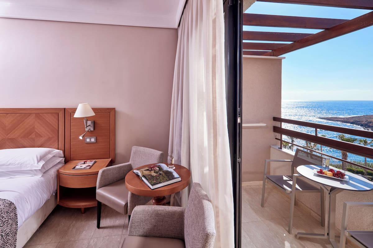 A hotel room with a bed, a nightstand, a lamp, a small table with a magazine, a chair, and a balcony with a table and fruit.