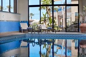 An indoor pool with clear blue water, lounge chairs, and a view of palm trees and buildings outside. - Atlantica Golden Beach -Adults Only (Kissonerga)