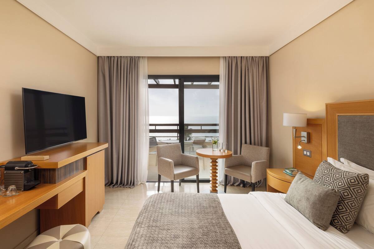 A hotel room with a bed, a desk with a TV, a chair, a small table with a lamp, and a view of the outdoors.