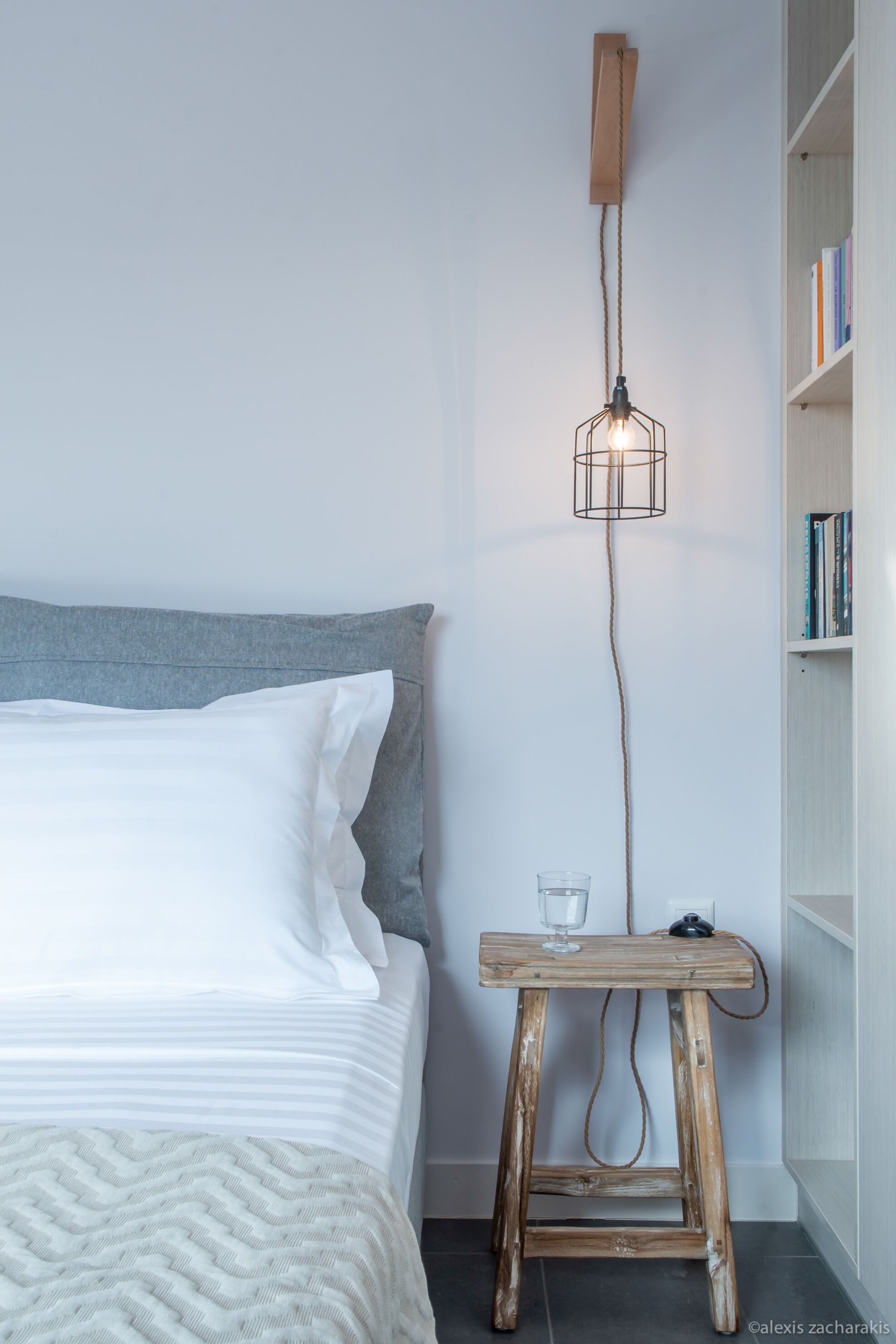 Un lit bien fait, avec du linge de lit blanc, une tête de lit grise et une table de chevet en bois sur laquelle se trouvent un verre et un livre. Un luminaire mural original est suspendu au-dessus du lit.