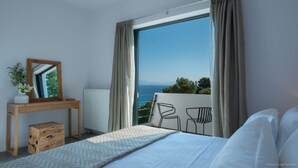 A bedroom with a large window, a bed, a bedside table with a mirror, and a view of the sea.