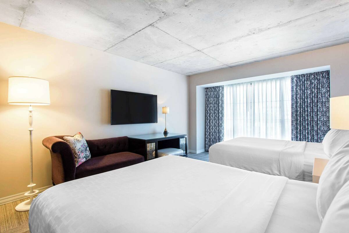 Habitación de hotel con dos camas, un televisor de pantalla plana montado en la pared, un escritorio con lámpara y un ventanal con cortinas.