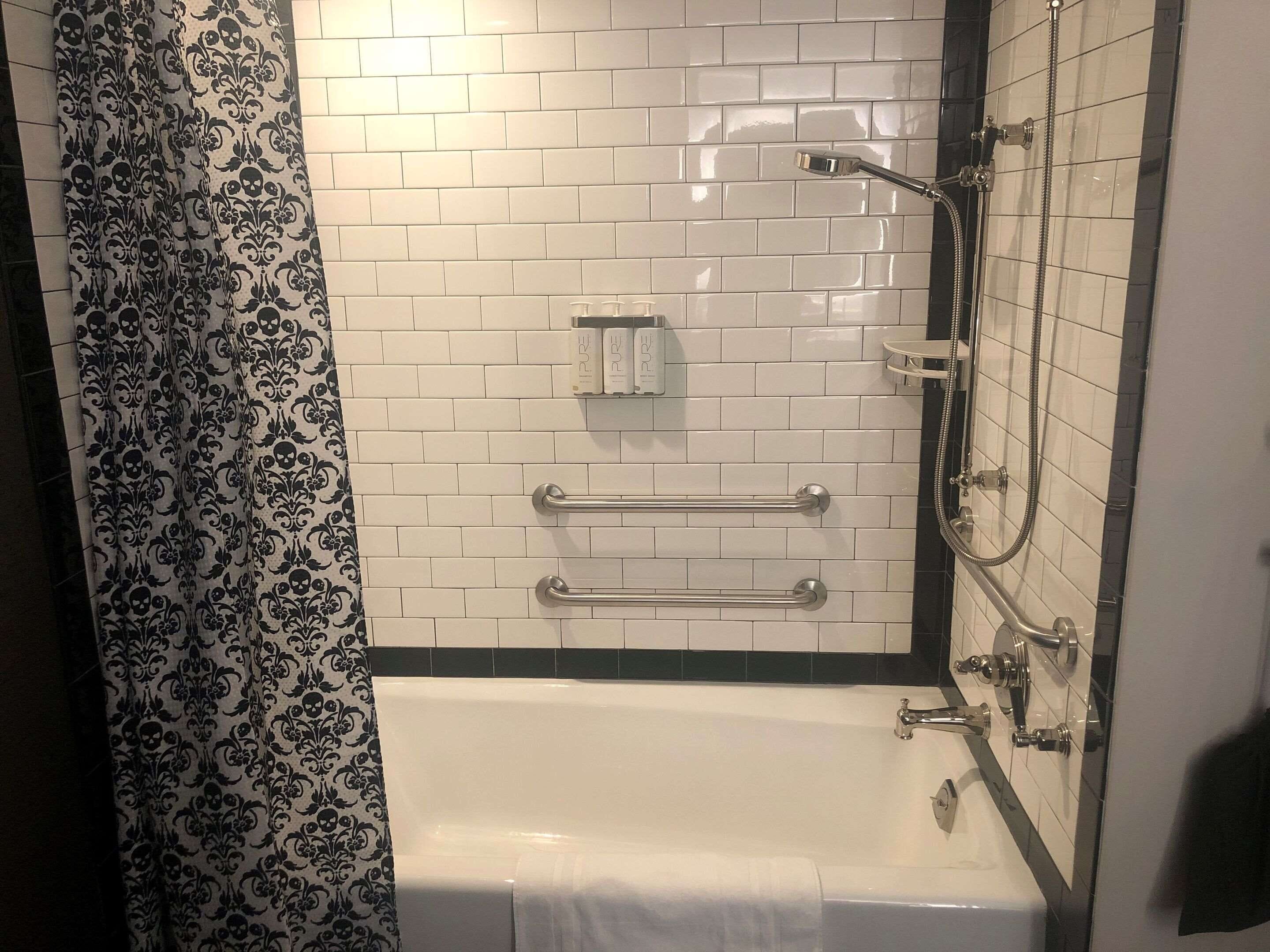 Un baño con paredes de azulejos blancos, una bañera, una ducha con cabezal de ducha manual y un perchero para toallas.