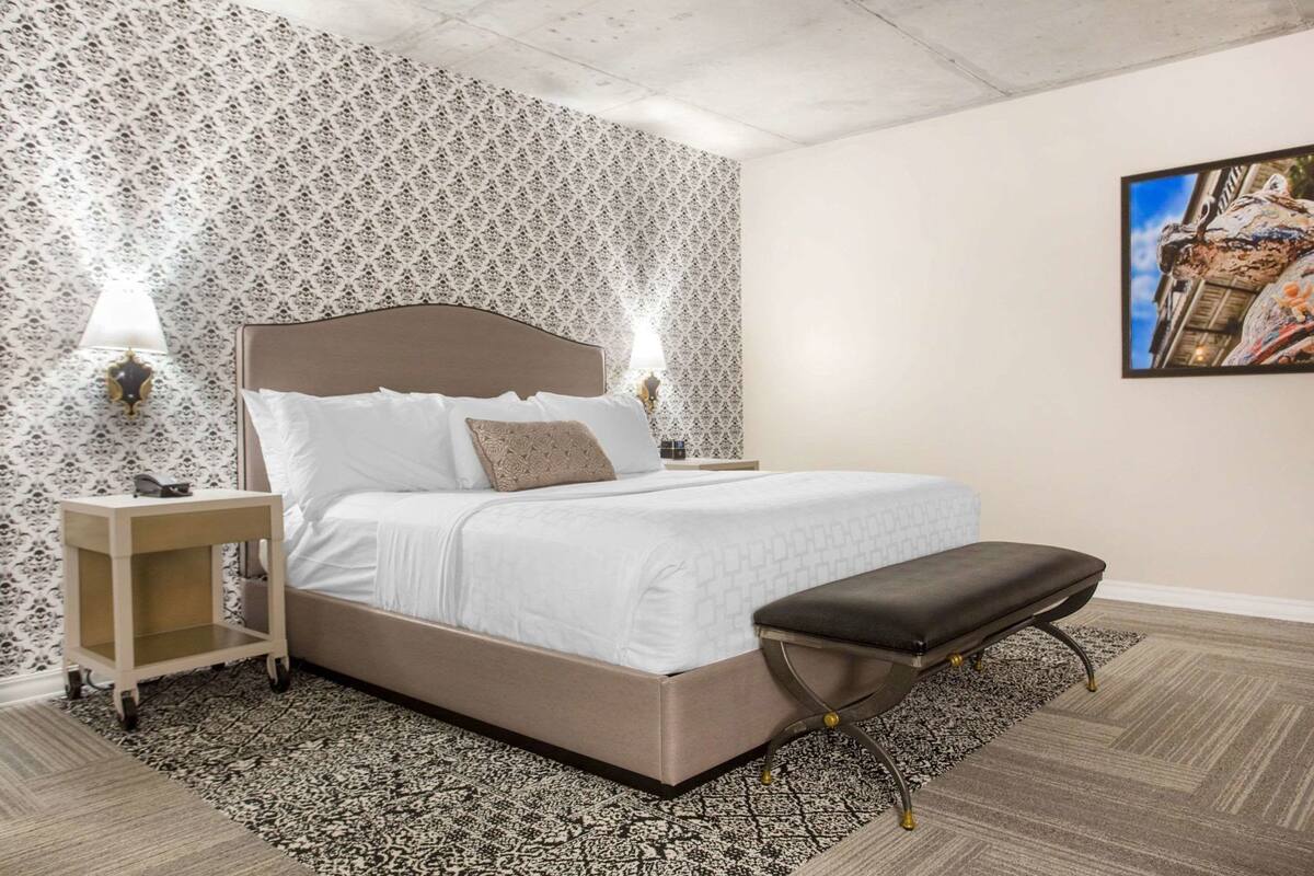 Un dormitorio con una cama, mesitas de noche, un banco, papel tapiz estampado y un cuadro en la pared.