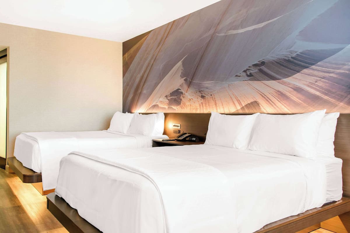 A hotel room with two beds, a large abstract mural on the wall, and a bedside table with a lamp.