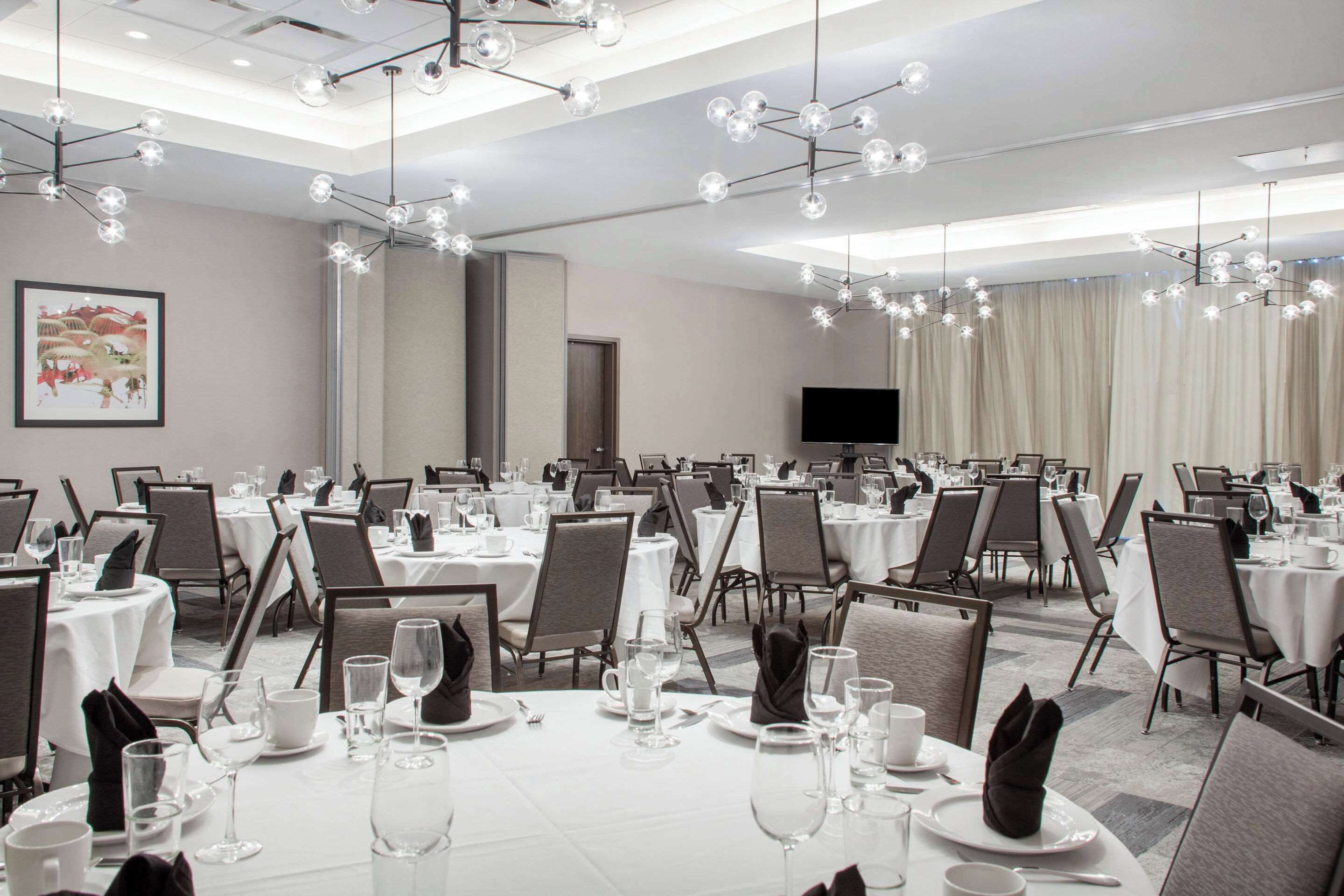 Une salle de banquet avec des tables rondes dressées pour le repas, un grand téléviseur à écran plat accroché au mur et des lustres modernes.