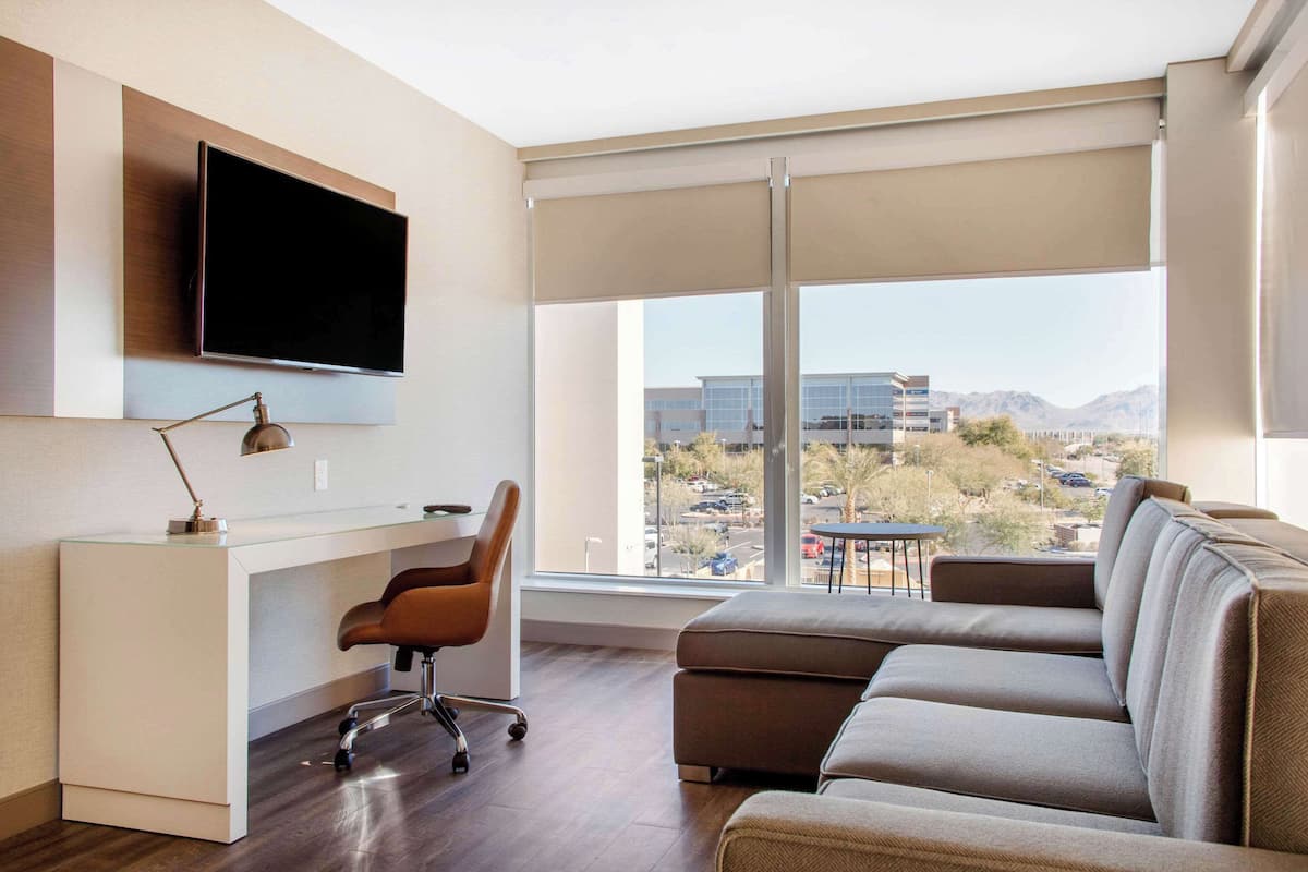 A modern living room with a flat-screen TV mounted on the wall, a glass desk with a chair, and a large window offering a view of a cityscape and mountains.