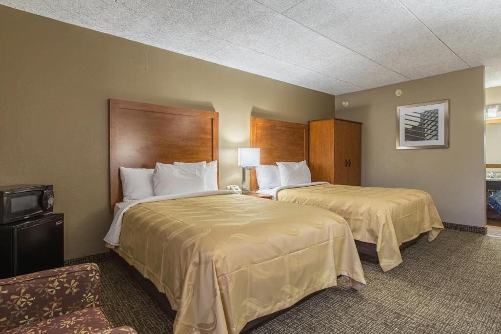 A hotel room with two beds, a desk, a chair, and a microwave.