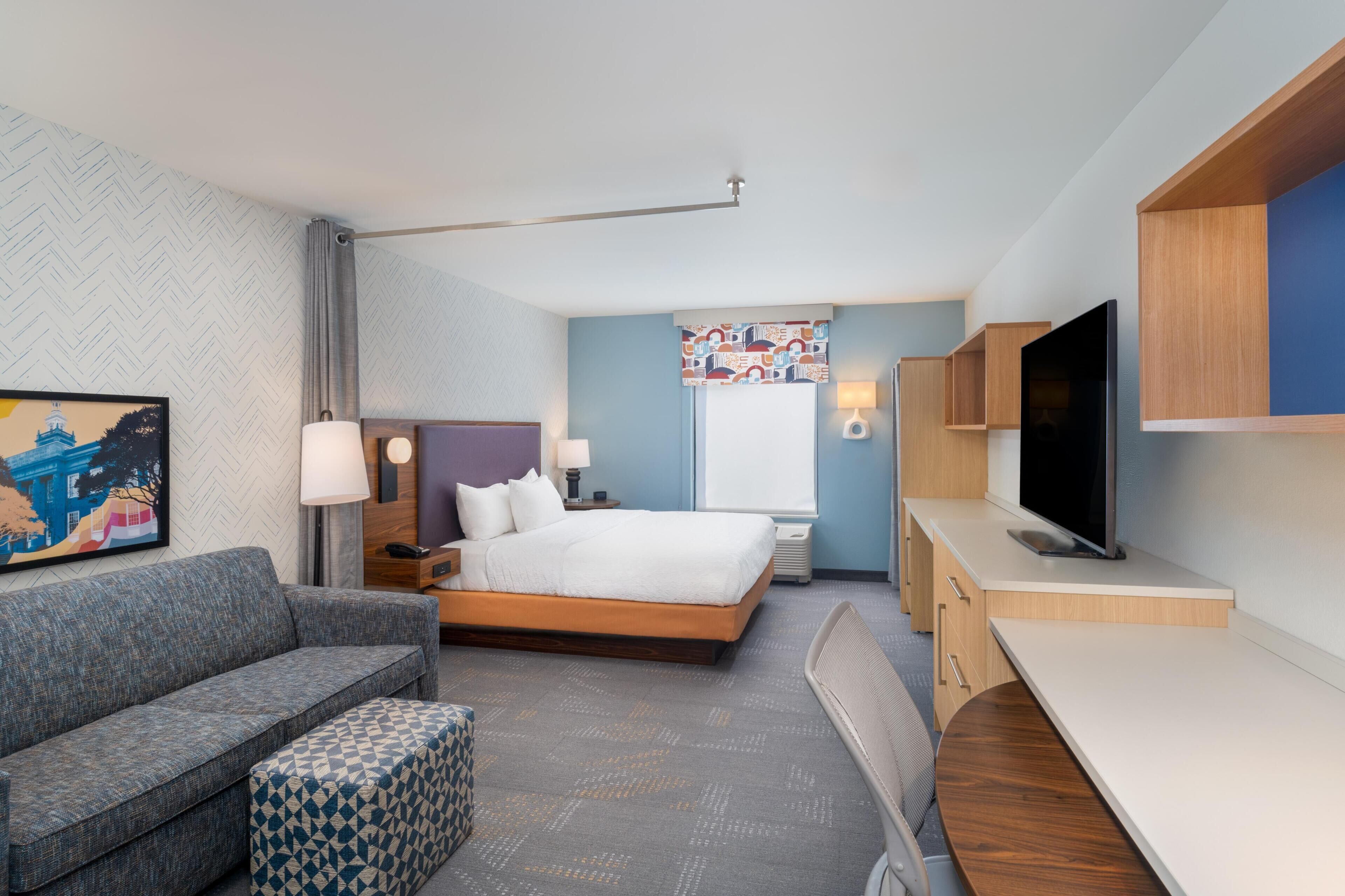 A modern hotel room with a bed, a sofa, a desk, a TV, and a painting on the wall.