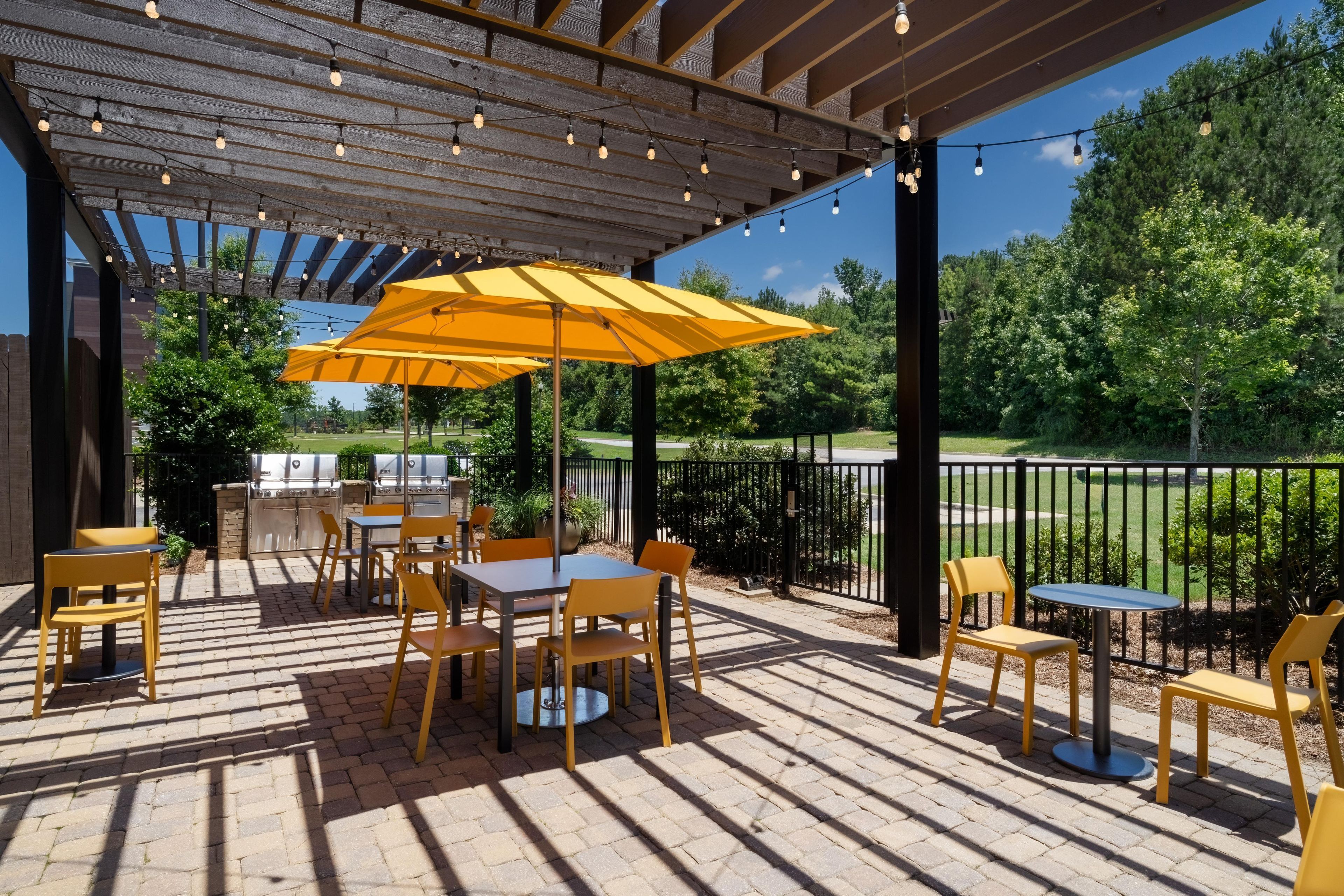 Une terrasse extérieure avec un sol en briques, des chaises jaunes, un grand parasol jaune et des guirlandes lumineuses.