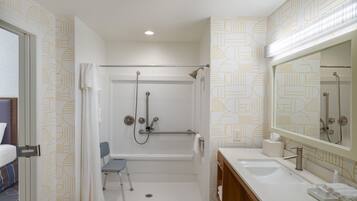 Baño con ducha, lavamanos, espejo y una silla para personas en silla de ruedas.