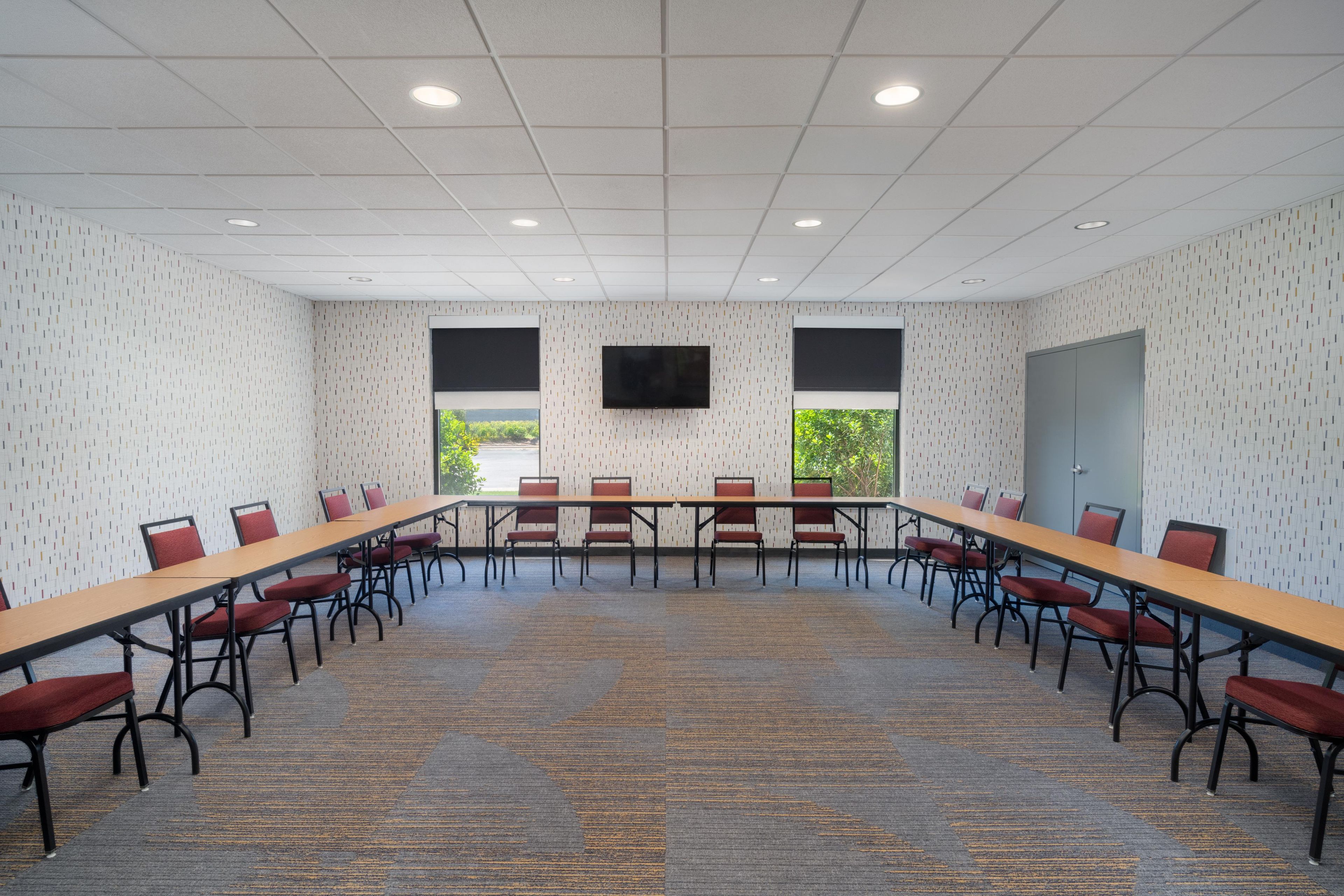 Uma sala de conferências com uma mesa comprida, cadeiras vermelhas, uma televisão de tela plana e janelas com vista para áreas verdes.