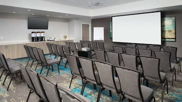 A conference room with rows of chairs, a projector screen, a flat-screen TV, and a coffee station.