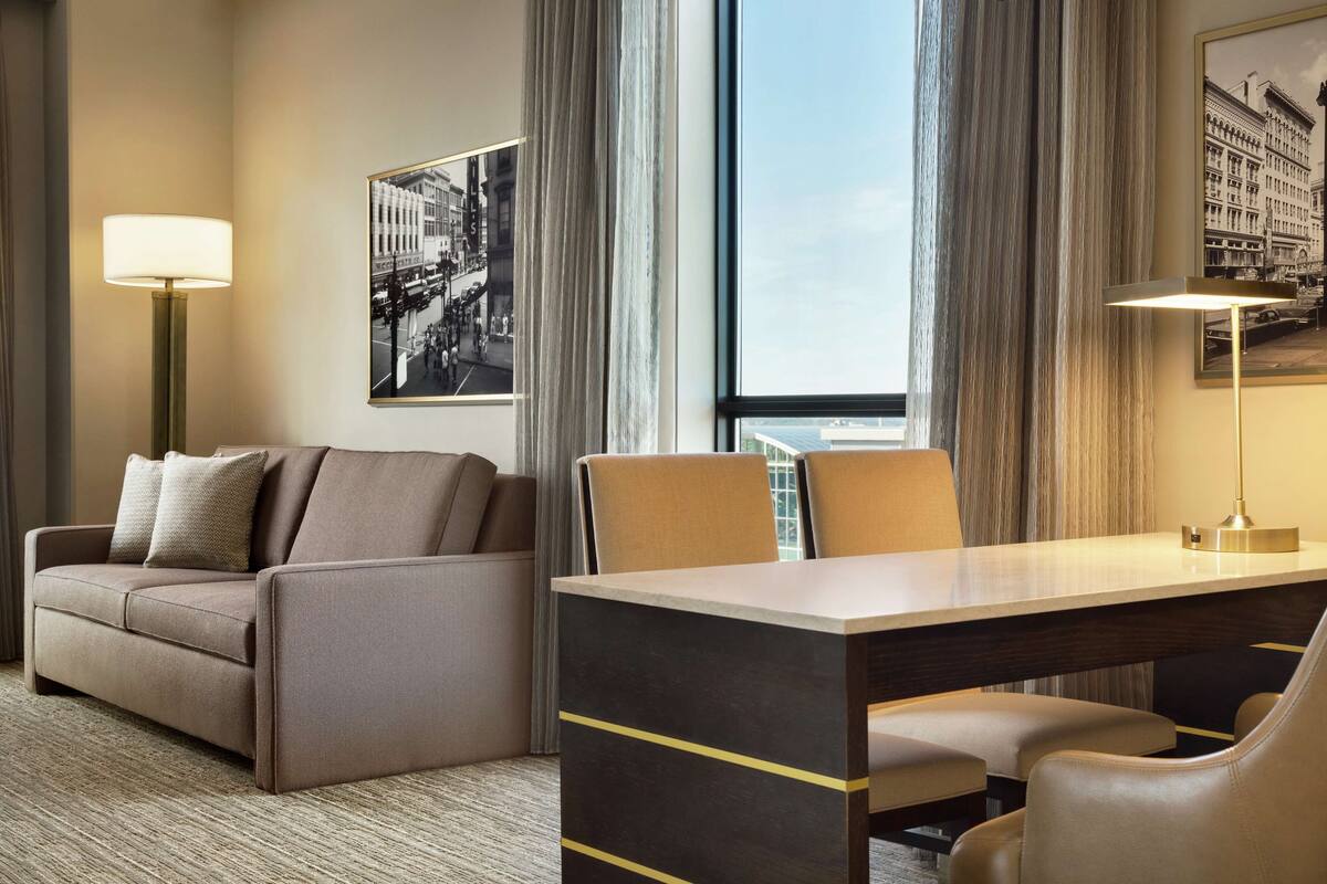 Een hotelkamer met een bank, een bureau, twee stoelen, een lamp en een groot raam met gordijnen.