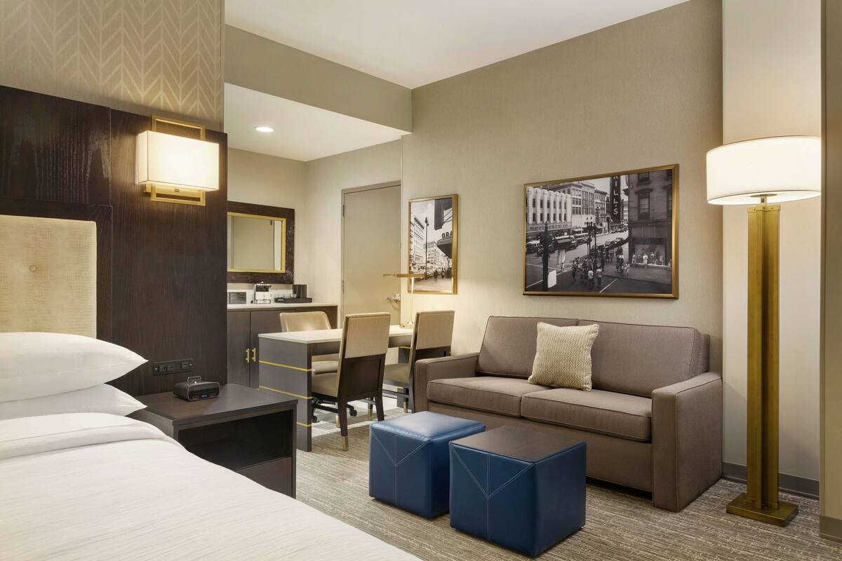 Een hotelkamer met een bed, een slaapbank, een bureau en een stoel. Er is een lamp, een foto aan de muur en een raam.