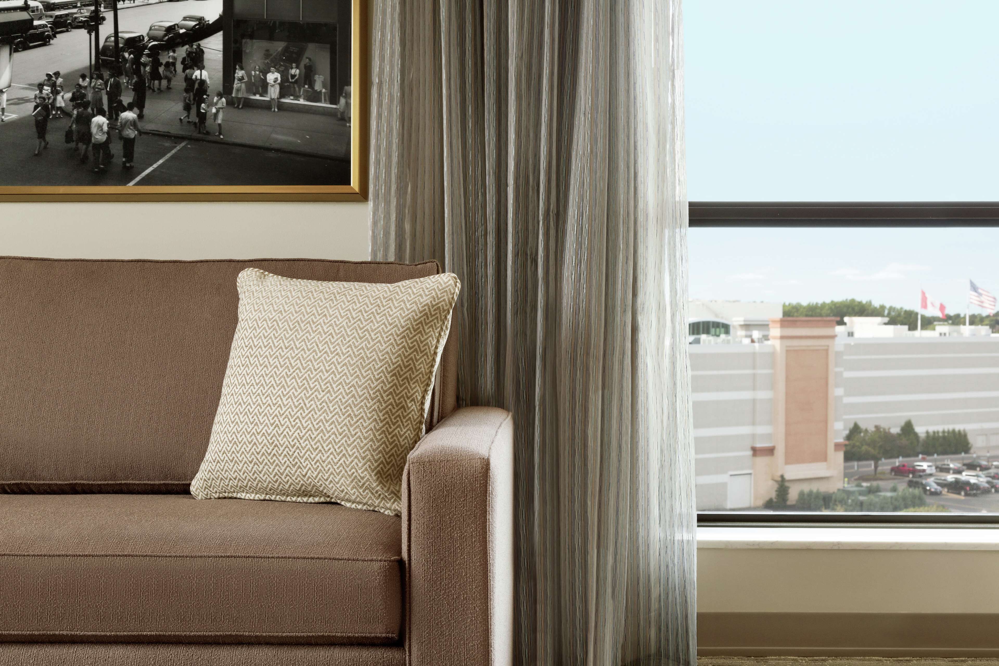 A brown sofa with a patterned pillow, a framed picture on the wall, and a view of a parking lot through a window with curtains.