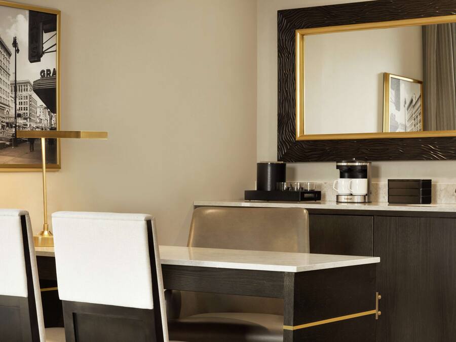 A modern hotel room with a dining area featuring a table and chairs, a coffee maker, and a framed cityscape picture on the wall.