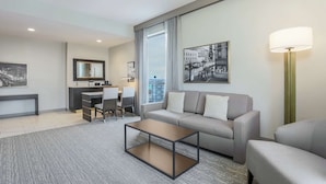 Een moderne hotelkamer met een grijze bank, een klein bijzettafeltje, een bureau met stoel, een groot raam met gordijnen en uitzicht op de stad.